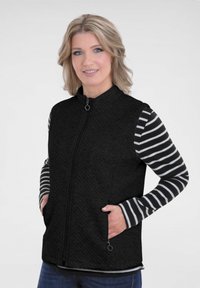 Zwarte vest met een gestructureerde afwerking, voorzien van een hoge kraag, ritssluiting en zijzakken. Daaronder is een gestreept overhemd met lange mouwen zichtbaar.