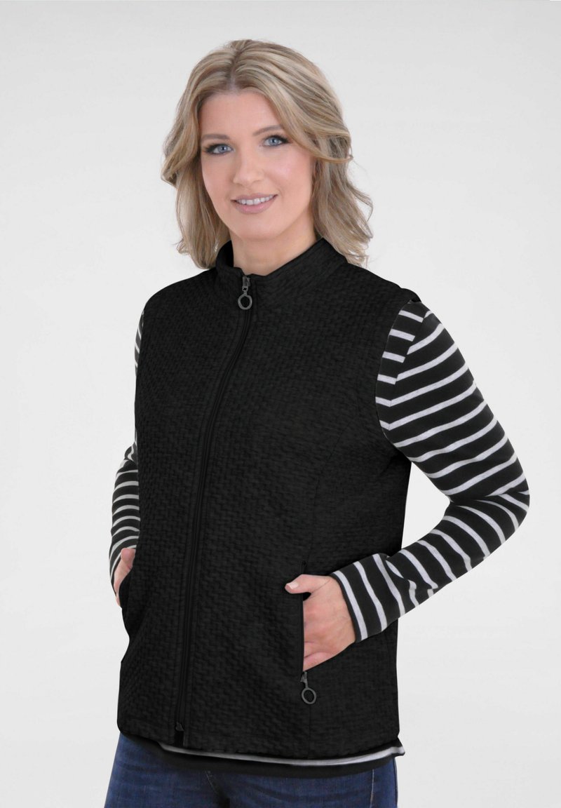 Zwarte vest met een gestructureerde afwerking, voorzien van een hoge kraag, ritssluiting en zijzakken. Daaronder is een gestreept overhemd met lange mouwen zichtbaar.