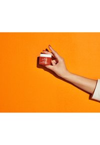 Pot de crème orange avec couvercle blanc, étiqueté "GINZING" par ORIGINS, tenu devant un fond texturé orange vif. Main et ongles visibles.