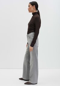 Vrouw staat zijwaarts gekeerd in een bruine coltrui, lichtgrijze wijde jeans en puntige donkerbruine schoenen tegen een effen witte achtergrond.