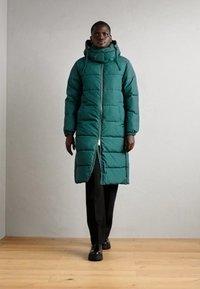 Dlhý, nadýchaný parkas v tmavozelenej farbe s vysokým golierom, predným zipsom a bočnými vreckami. Tkanina sa zdá byť hladká s prešívanými vzormi.