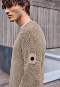 Beige gebreide trui met structuur, voorzien van een ronde hals en een patch met een knoopdetail op de linkermouw.