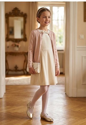 Niña joven con vestido crema, cárdigan rosa, medias blancas y zapatos dorados, sosteniendo un bolso blanco, de pie en una habitación luminosa con suelo de madera.