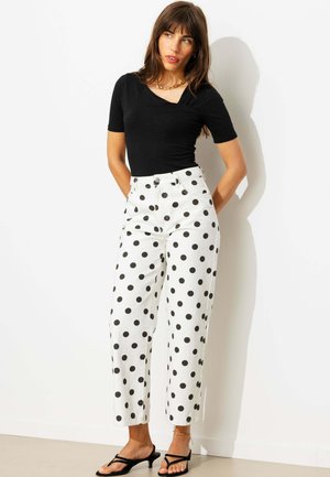 Femme portant un haut noir à manches courtes, un pantalon blanc à gros pois noirs, des sandales noires à brides et un collier en or, debout contre un mur blanc.