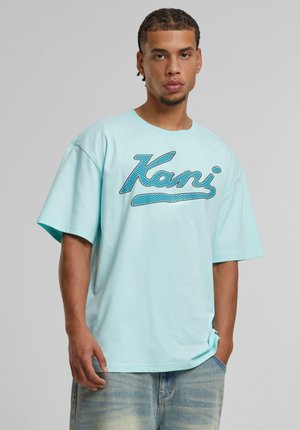 Camiseta de algodón oversize azul celeste, con un prominente logo azul "Kani" impreso en el frente. El modelo lleva puestos unos pantalones cortos de mezclilla claros.