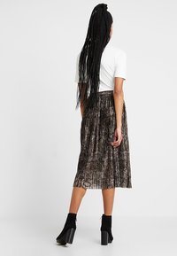 Mujer con cabello trenzado largo, vistiendo una blusa blanca de manga corta, una falda midi con estampado de serpiente plisada y botas de tacón negras, de pie, de espaldas.