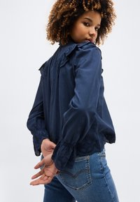 Νεαρή γυναίκα με σγουρά μαλλιά, ντυμένη με μπλε μπλούζα μακριάς μανίκας με δαντέλα και μπλε τζιν, κοιτάζοντας πάνω από τον ώμο της.