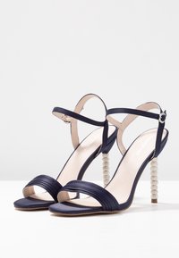 Námořnické saténové sandály na vysokém podpatku s křížovými pásky, otevřenou špičkou a průhledným podpatkem zdobeným krystaly. Hladká textura a minimalistický design.