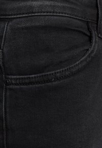 Vaqueros de mezclilla negra con un bolsillo frontal con bordes cosidos y un pequeño detalle de botón, mostrando una tela lisa y con textura.