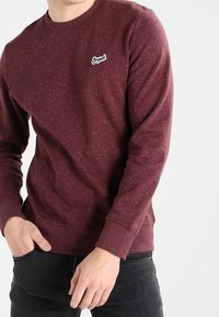 Muž v vínovém melírovaném svetru s dlouhým rukávem a malým logem "Original" na levé straně hrudi a tmavých kalhotách.