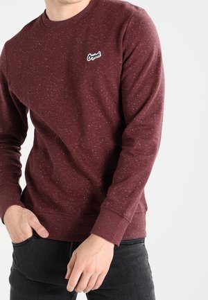Uomo che indossa un pullover a maniche lunghe di colore marsala con un piccolo logo "Original" sul lato sinistro del petto e pantaloni scuri.
