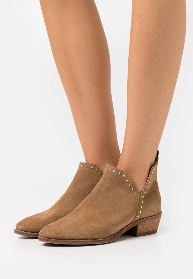 Bottes en suède beige montant jusqu'aux chevilles, avec des talons empilés bas et de petites clous métalliques le long des bords supérieurs, portées sur des jambes nues.