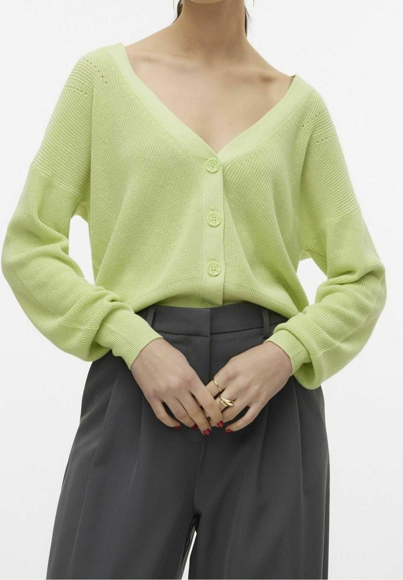 Cárdigan de punto verde lima con escote en V, cierre de cinco botones y mangas holgadas, combinado con pantalones de tiro alto grises.