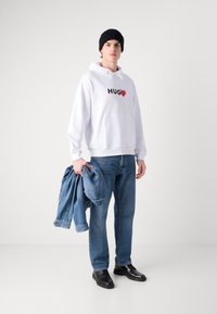 Felpa bianca con grafica "HUG" e cuore, abbinata a jeans in denim blu e giacca di denim tenuta in mano. Un berretto nero e scarpe completano il look.