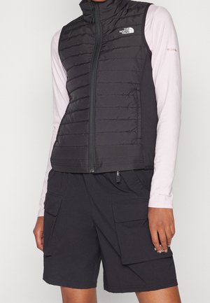 Person iført en sort quiltet North Face vest, lyserød langærmet skjorte og sorte cargoshorts med store lommer.