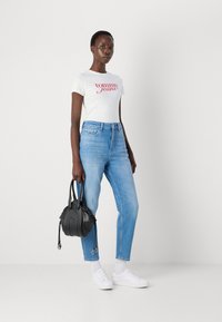 Bela majica s rdečim logotipom "Tommy Jeans", svetlo modri hlače z visokim pasom, črna torbica in bele superge. Minimalističen, sproščen outfit.