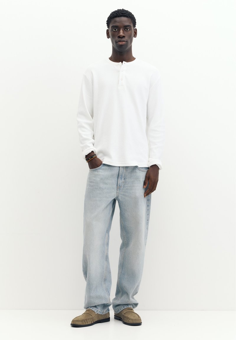 Jeune homme debout les mains dans les poches, portant une chemise blanche à manches longues, un jean bleu clair et des mocassins beiges sur un fond blanc uni.