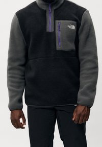 Sort og grå fleece-pullover med en kvart lynlås, en lilla lynlås-træk og en brystlomme med et logo-patch. Struktureret stof.