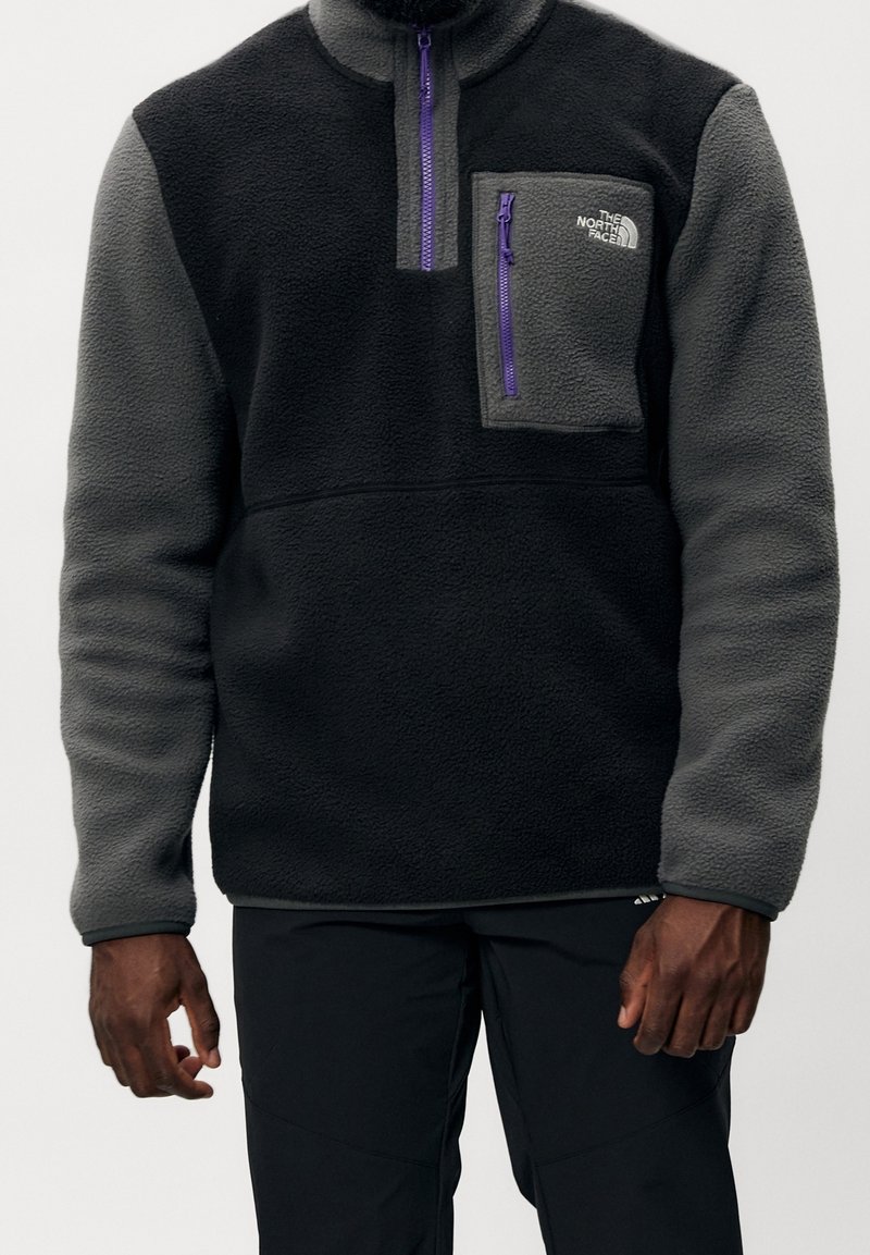 Sort og grå fleece-pullover med en kvart lynlås, en lilla lynlås-træk og en brystlomme med et logo-patch. Struktureret stof.