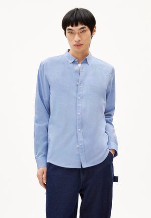 Light blue long-sleeve button-up shirt in woven fabric, with a modern collar and slightly rounded hem, paired with dark denim trousers.