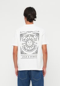 T-shirt blanc à manches courtes présentant un design graphique noir d'un soleil et du texte au dos. Le matériau semble être en coton ; coupe classique.
