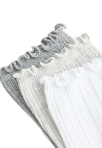 Trois paires de chaussettes courtes en maille côtelée en blanc, gris clair et gris foncé avec bords à volants empilées en diagonale.