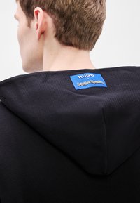 Μαύρο φούτερ με υφασμάτινη υφή, που έχει μια μπλε ετικέτα στην πλάτη με τις λέξεις «HUGO ×» και «Jujutsu Kaisen» σε χρυσά γράμματα.