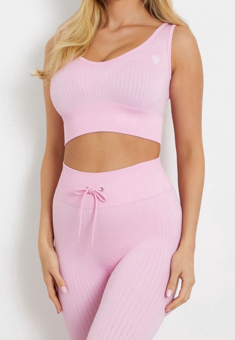 Kobieta ubrana w dopasowany różowy stanik sportowy i legginsy z wysokim stanem, z regulowanym sznurkiem w legginsach, stojąca na tle białym.