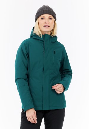 Vrouw in een teal kapwinterjacket en een donkergrijze gebreide beanie, staand tegen een witte achtergrond.