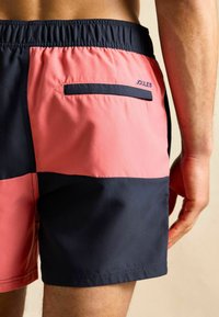 Różowo-granatowe kąpielówki w blokach kolorystycznych z tylną kieszenią. Posiadają elastyczny pas i widoczne logo. Gładka tekstura.