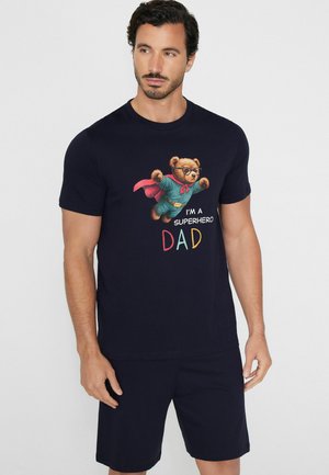 Mężczyzna w czarnym t-shircie i szortach, na t-shircie nadruk latającego misia z peleryną i napis „Jestem superbohaterem tatą.”