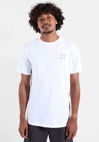 T-shirt en coton blanc avec un col rond et des manches courtes, arborant un petit motif floral violet sur la poitrine gauche.