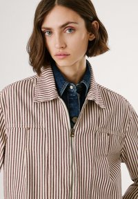 Giacca a strisce con un motivo bordeaux e crema, presenta una chiusura con zip, un colletto e una tasca sul petto, indossata sopra una camicia di jeans.