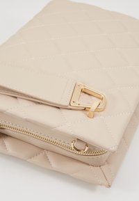 Borsa a tracolla beige trapuntata realizzata in materiale sintetico. Presenta dettagli in oro, chiusura con zip e una tracolla removibile.