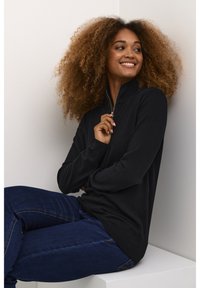 Juodas zip-up megztinis su raštuotu apykakle, derinamas su tamsiai mėlynomis džinsomis. Megztinis turi elegantišką dizainą ir patogią prigludimą.