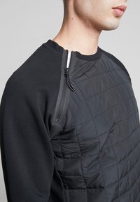 Svart sweatshirt med vadderad front, med en dragkedja på axeln. Tillverkad av en blandning av släta och texturerade material.