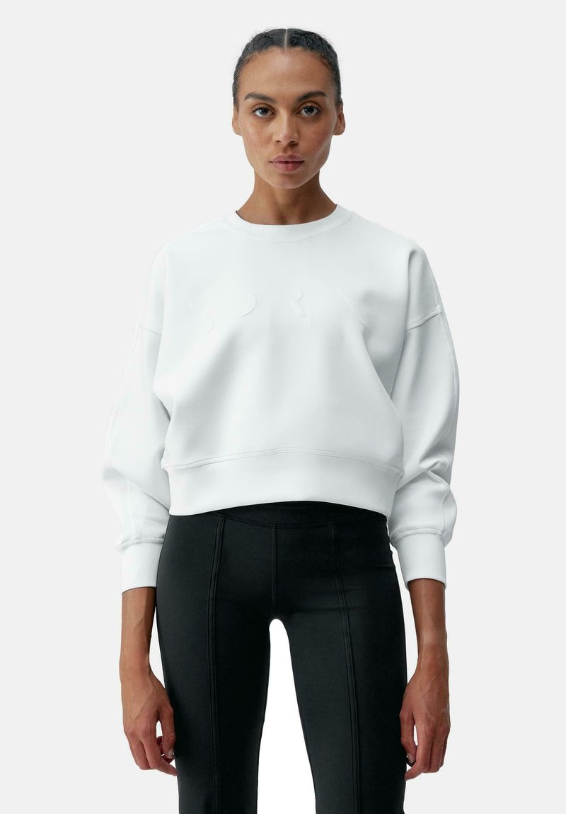 Sweatshirt en coton mélangé blanc avec un col rond, des épaules tombantes et une inscription en relief subtile. Associé à un legging noir avec des détails de couture.