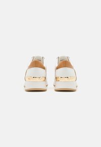 Balti ir tan odiniai sportbačiai su bežiniu padu ir auksiniu akcentu ant kulno, su metalinėmis ženklais ir tekstūruotu detalėmis.