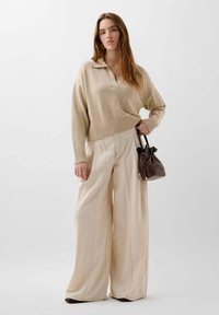 Beige gestrickter Pullover mit Kragen, kombiniert mit cremefarbenen weit geschnittenen Hosen. Hält eine kleine braune Lederhandtasche mit Zugbandverschluss.