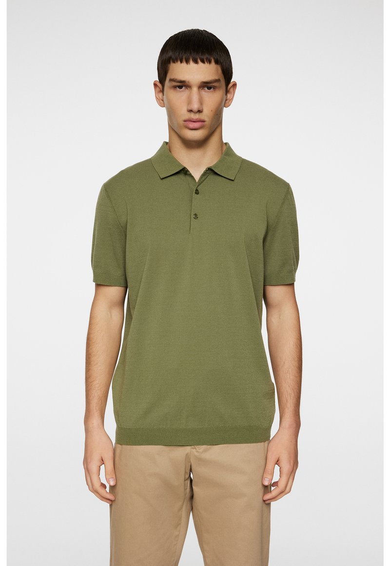 Polo shirt en tricot vert olive avec un col, une patte de boutonnage à trois boutons et des manches courtes, associé à un pantalon beige clair. Texture lisse.