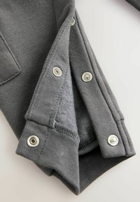 Szary materiał bluzy z miękką podszewką, z metalowymi zatrzaskami na mankietach i teksturowaną zewnętrzną powierzchnią.