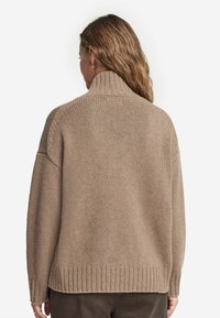 Beige gestrickter Pullover mit hohem Kragen, überschnittenen Schultern und Rippbund. Die Rückansicht zeigt eine lockere Passform und eine weiche Textur.