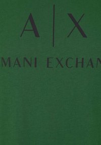 Zelena bombažna majica z črnim tiskanim besedilom "A|X" in "ARMANI EXCHANGE." Tkanina ima gladko teksturo, brez dodatnih vzorcev.