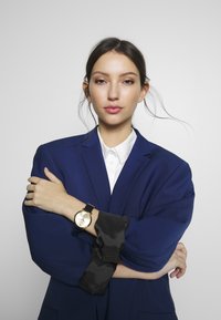 Gul armbandsur med rund urtavla, svart läderband, bärs med en mörkblå blazer och en vit skjorta. Släta tygaccentuer synliga.