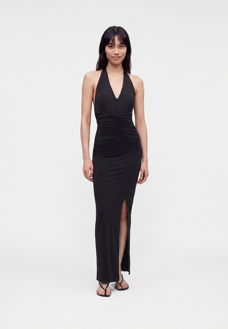 Γυναίκα που φοράει μαύρο αμάνικο φόρεμα με halter neck, πιέτες και σκίσιμο μπροστά, στέκεται μπροστά σε απλό λευκό φόντο.