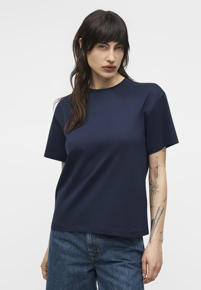 Γυναίκα με σκούρα μαλλιά, ντυμένη με μπλε σκούρο μπλουζάκι και μπλε τζιν, που δείχνει τα τατουάζ στο αριστερό της αντιβράχιο, στέκεται μπροστά σε απλό φόντο.