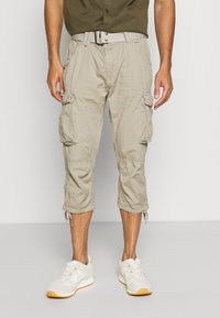 Pantalons cargo beiges avec plusieurs poches latérales, longueur genoux, poignets ajustables et une ceinture texturée. Portés avec des baskets blanches.
