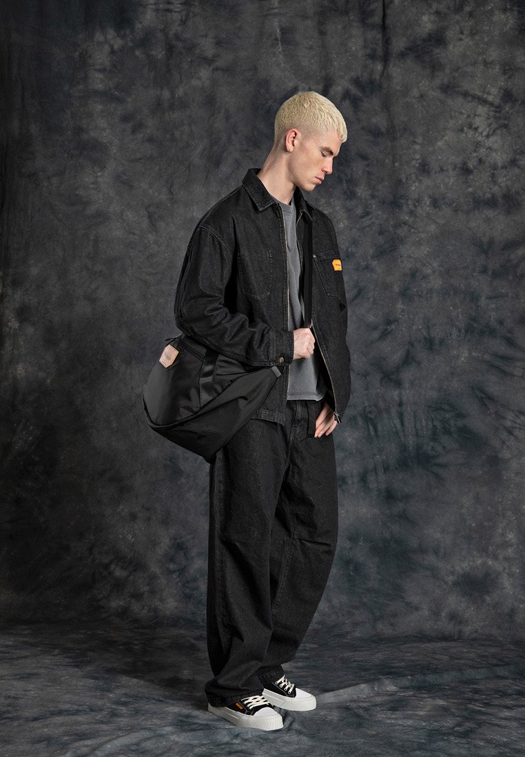 Zwarte denim jacket en broek, grijze top, zwarte tas, witte sneakers met zwarte accenten. Model staat tegen een gestructureerde grijze achtergrond.