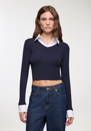 Top lungo a maniche lunghe cropped color navy con scollo a V, sovrapposto a una camicia bianca con colletto. Jeans in denim blu a vita alta con dettagli di cucitura visibili.