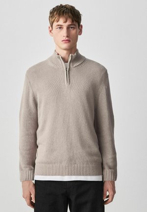 Junger Mann trägt einen hellbeigen Strickpullover mit halbem Reißverschluss über einem weißen Hemd und schwarzen Hosen, steht vor einem einfarbigen hellen Hintergrund.